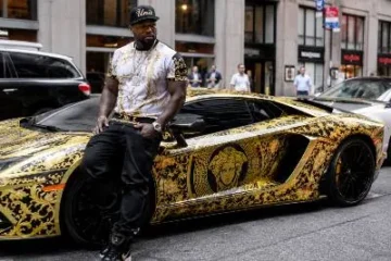 50 Cent also known as Curtis James Jackson III standing beside one of his luxury or exotic vehicles representing his car collection estimated at approximately 10.5 million dollars including a Ferrari Enzo worth 2.4 to 3 million dollars a Maserati MC12 one of only 50 road legal examples ever produced multiple Lamborghini Aventadors including a Versace wrapped Aventador S four Rolls-Royces maintained by DJ Envy a Bugatti Chiron and a Bugatti Veyron beginning from a 35000 dollar Toyota Land Cruiser purchased at age 18 in South Jamaica Queens New York